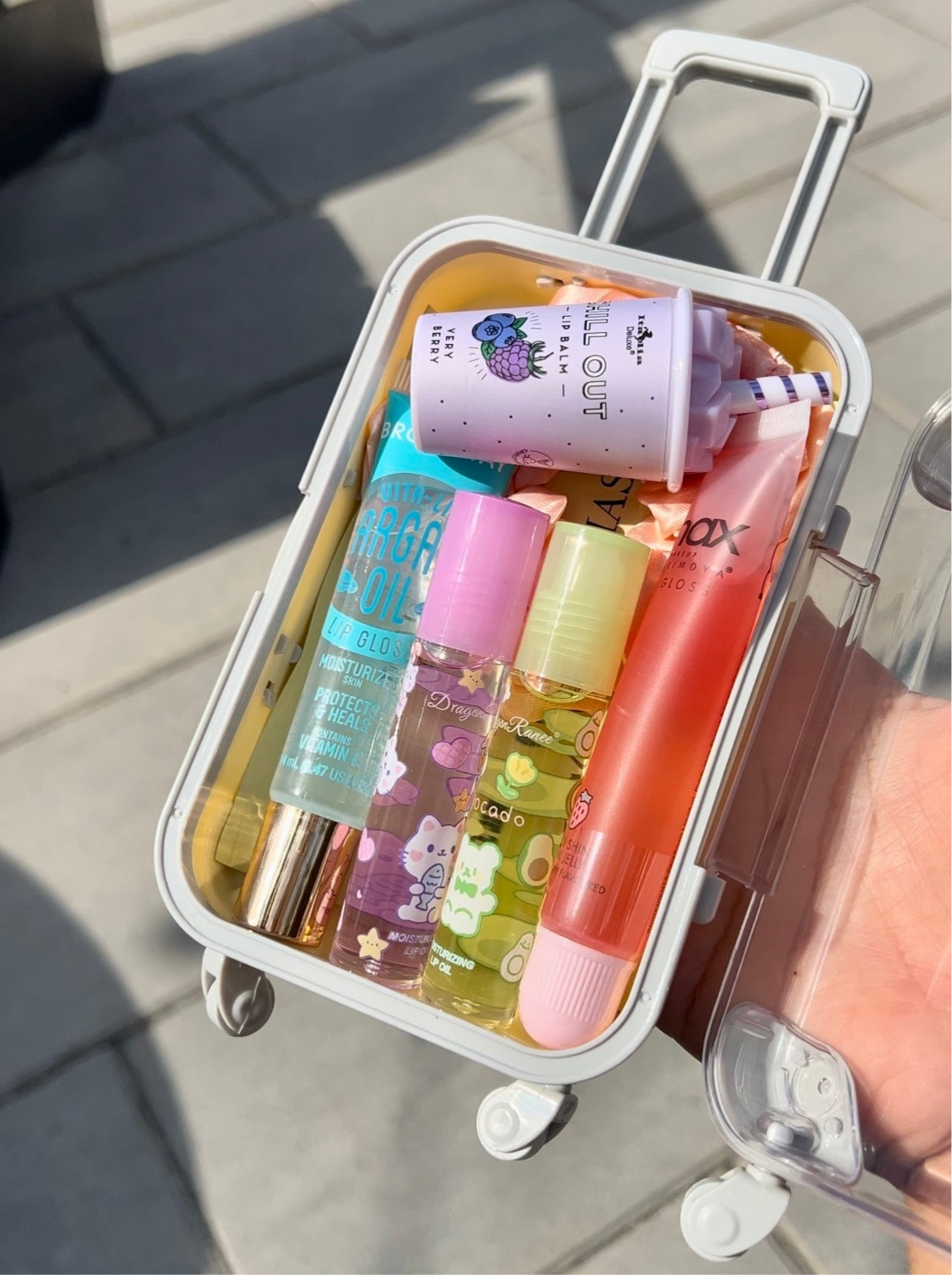 A mini suitcase-shaped container filled with various lip glosses, lip balms, and a scrunchie, all in different colors.