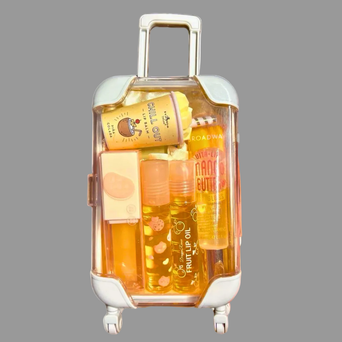 A clear rolling cosmetic orange suitcase with white trim and wheels, filled with assorted beauty products including lip oil, cuticle oil, and a lip balm, held by a hand against a plain white background.