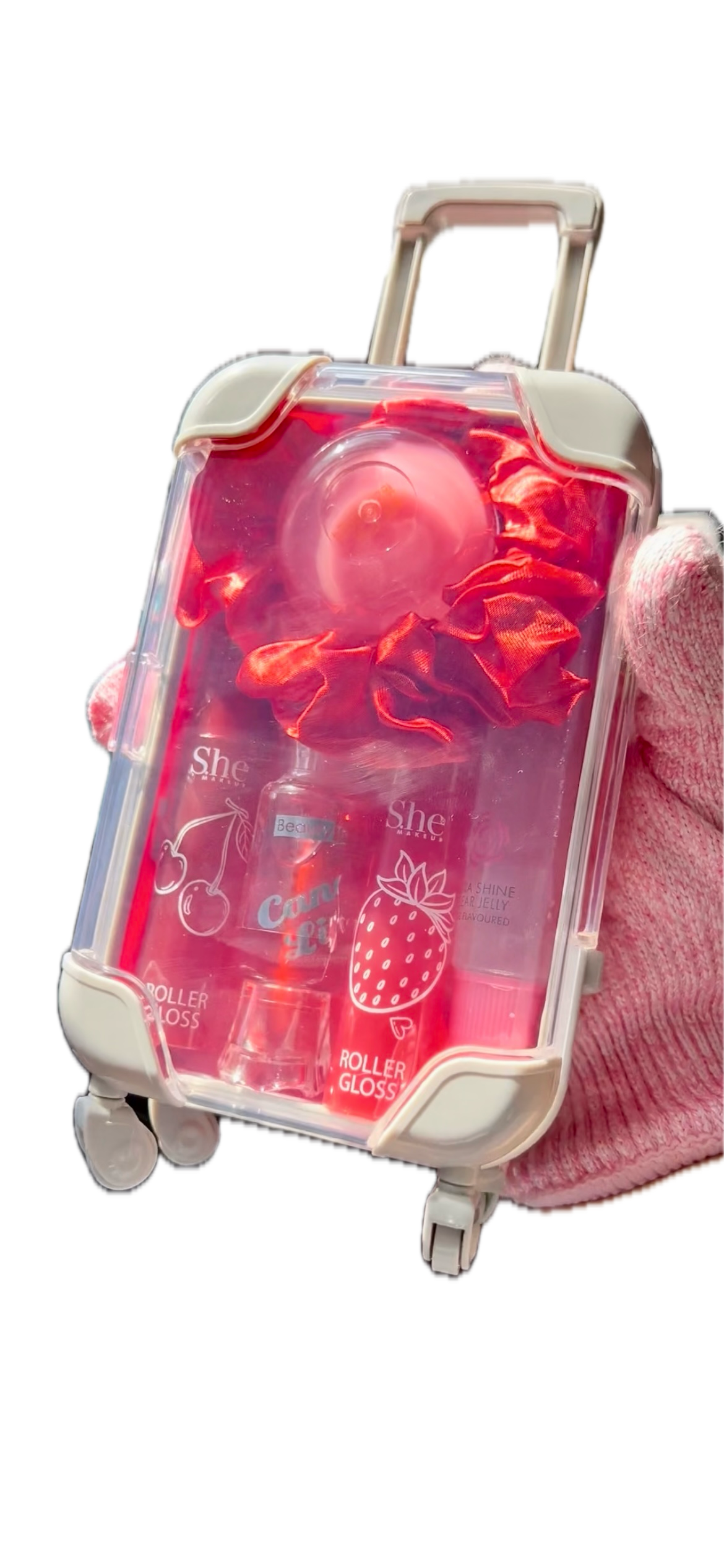 Dark pink textured knit sleeve holding a clear rolling suitcase filled with red rolled cosmetics, including a round pink item and bottles with strawberry and cherry graphics, isolated on a white background.