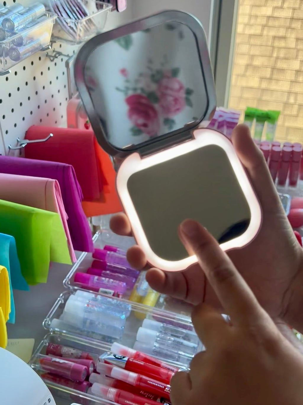 Hand holding a compact mirror with LED lights in front of colorful stationery items.